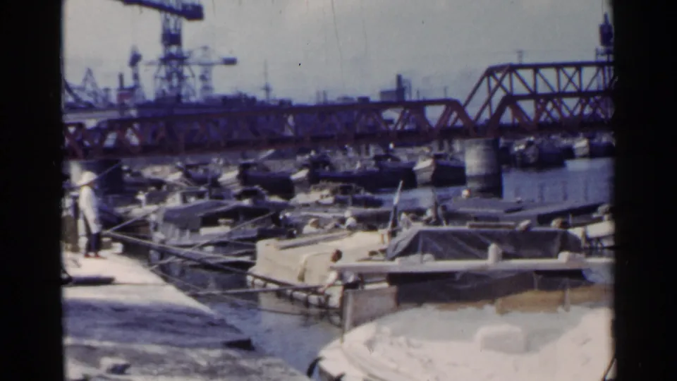 1953 Japan dock scene with workers near small boats, a red truss bridge over the water, cranes, and factory structures in the distance.