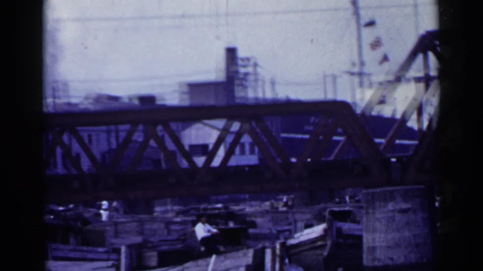 1953 Japan harbor frame showing the truss bridge closer in view, with boats below and cranes or factory structures beyond the span.