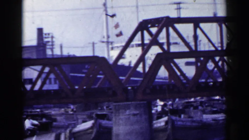 1953 Japan industrial harbor view with the red truss bridge filling the upper frame and small boats and a worker visible below.