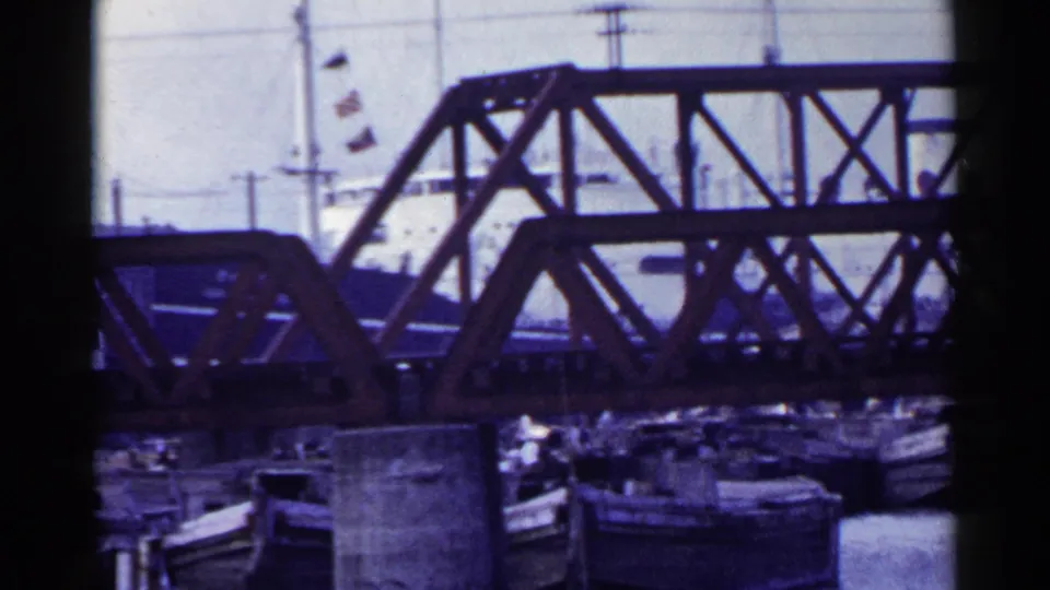 1953 Japan 8mm frame showing a steel truss bridge above moored boats, with overhead wires and flags in the background.