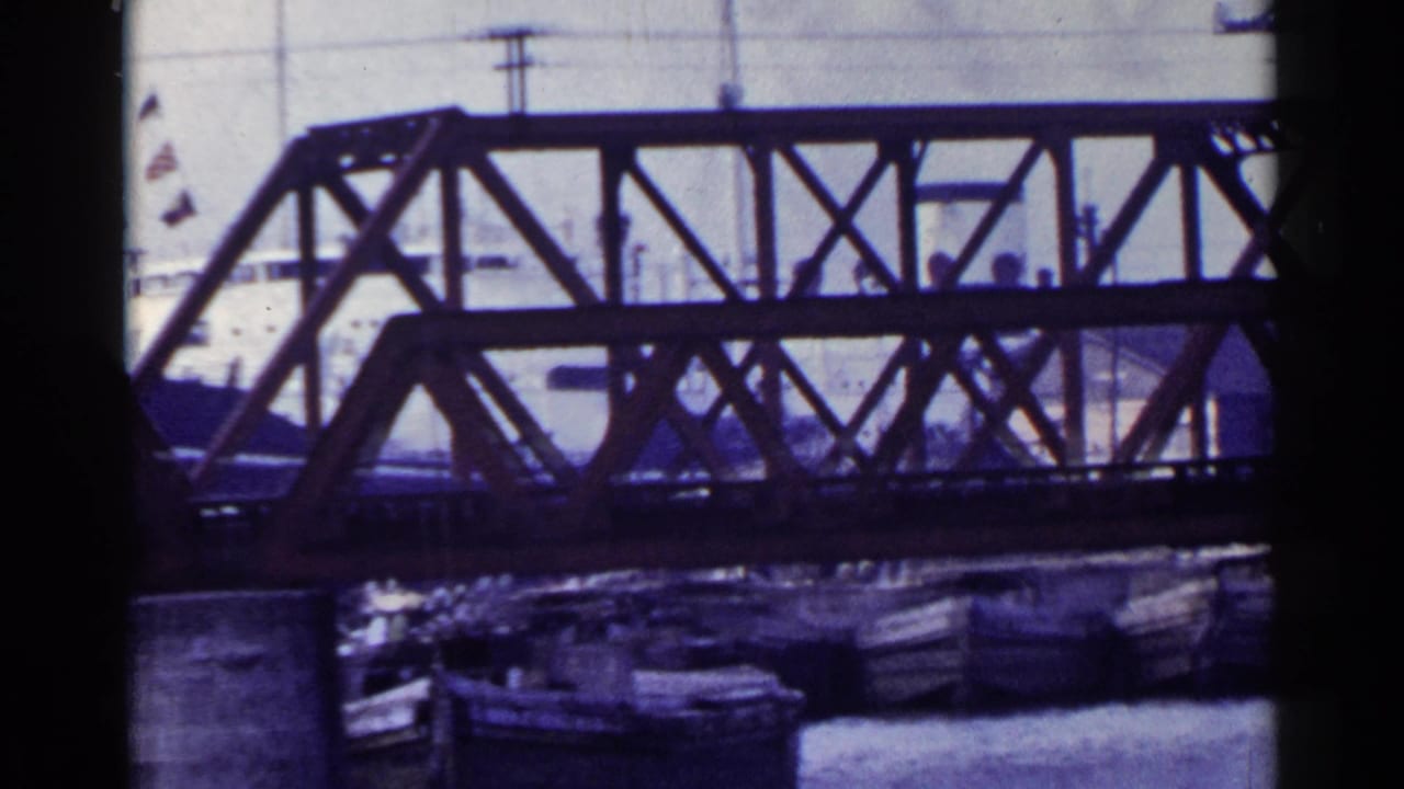 Steel truss bridge crossing a harbor channel with boats and flags behind it.