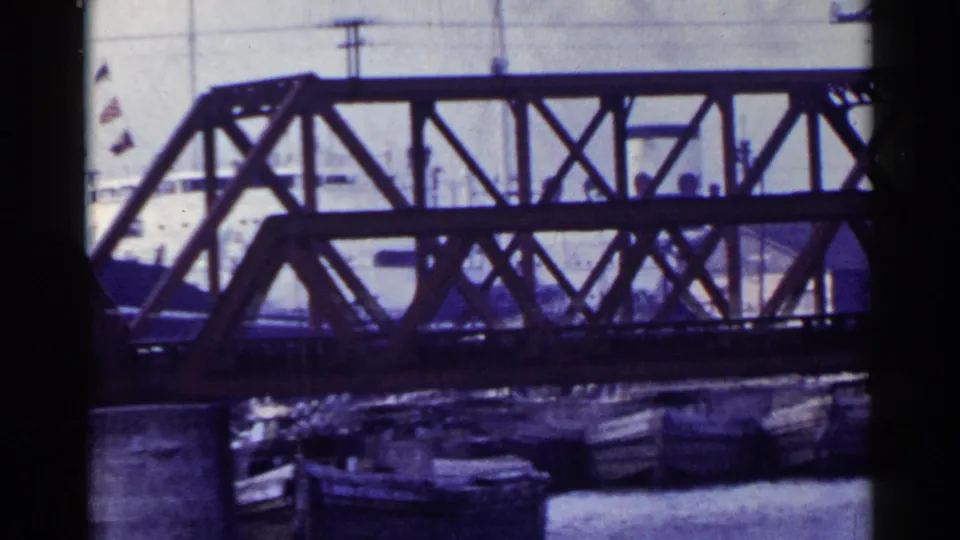 1953 Japan home-movie frame with a steel bridge, docked boats, utility poles, and flags partly visible beyond the span.