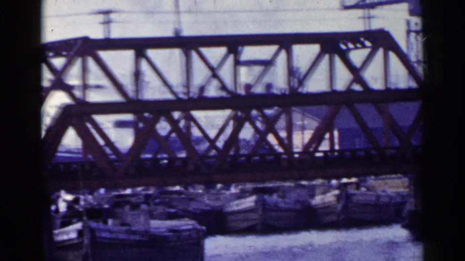 1953 Japan 8mm view of the bridge structure crossing above moored boats, with a pale cloudy sky and utility lines behind it.
