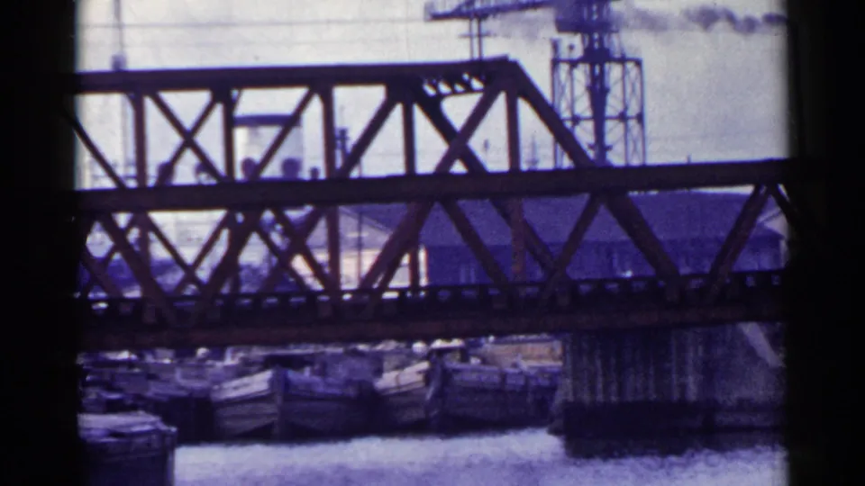 1953 Japan 8mm frame showing the steel truss bridge, moored boats, water surface, utility lines, and a large vessel or structure at right.