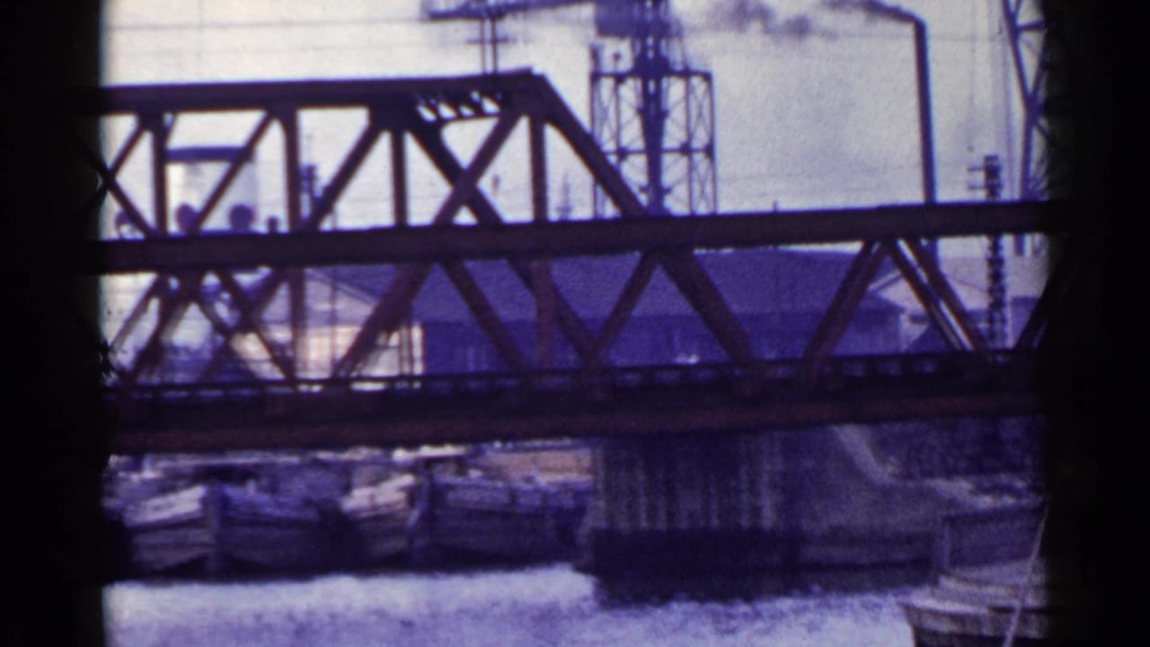 Steel truss bridge over a river with boats and industrial structures in the background.