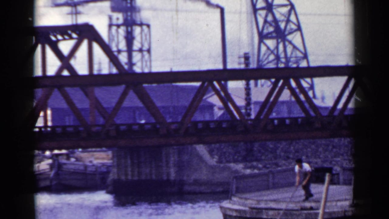 Man in a white shirt stands on a wooden barge beneath a steel truss bridge.