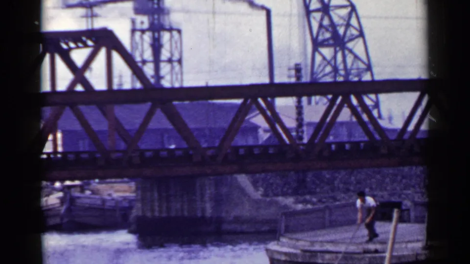 1953 Japan 8mm frame with an adult man in a white shirt standing on a wooden barge beneath a steel truss bridge.