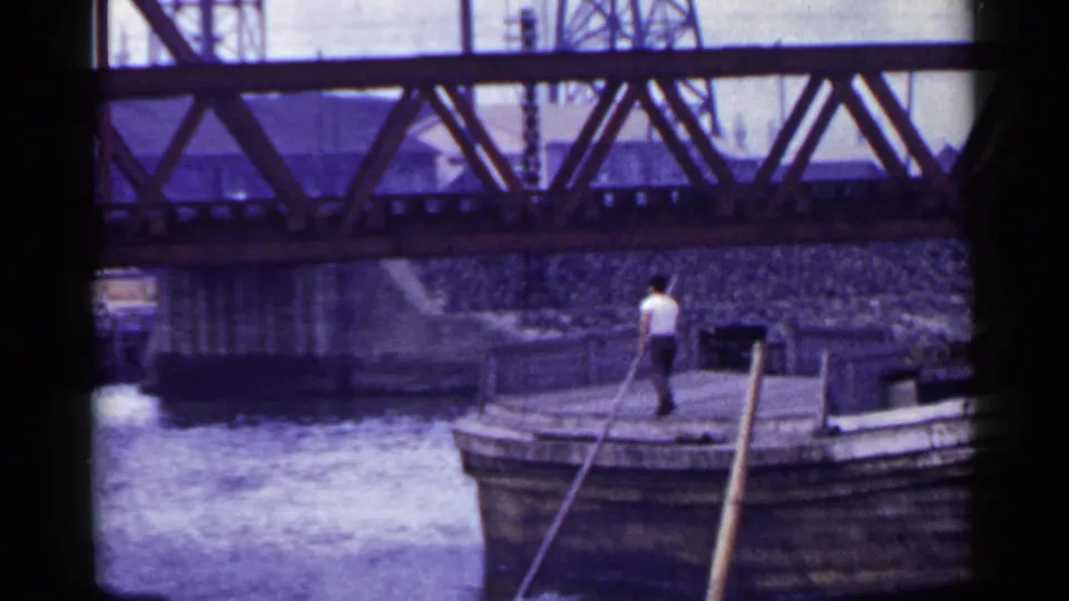 1953 Japan 8mm frame showing a man in a white shirt using a long wooden pole on a barge below a steel bridge.