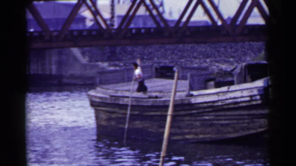 1953 Japan 8mm frame with a wooden barge in the foreground and a man handling a long pole beneath the bridge.