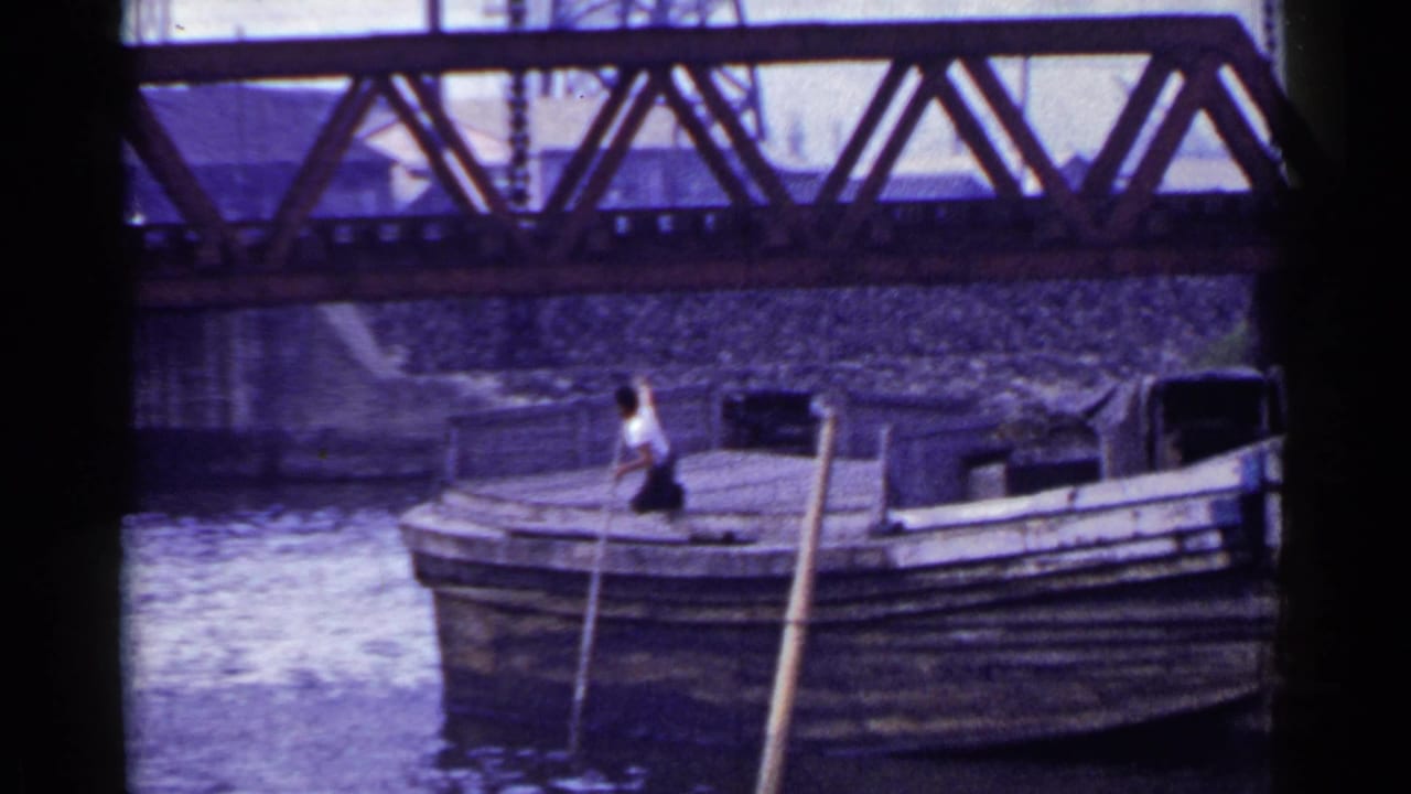 Man with a long pole stands on a wooden boat beneath a metal truss bridge.