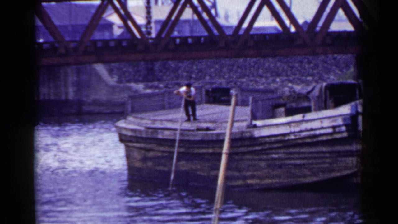 A man stands near the bow of a wooden boat with long poles in the water.