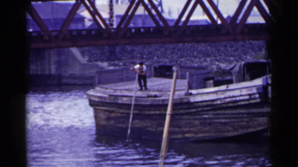 A distant 1953 Japan frame shows a man balancing on a wooden boat as long poles lean into the river near a bridge.