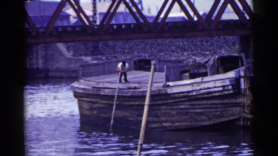 A man in a light shirt and dark trousers stands on a large wooden boat on a river in Japan, 1953.