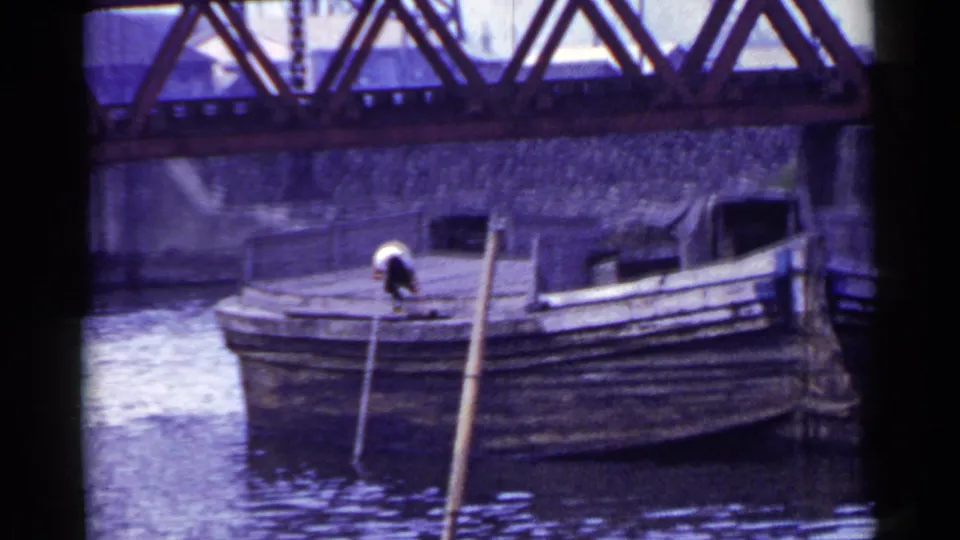 In this 1953 Japan frame, the boatman bends over on the wooden deck while poles lean beside the boat in the river.