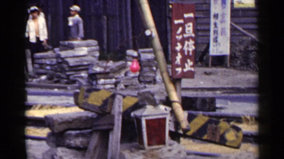Japanese street construction scene with stacked sandbags, roadside barriers, visible workers, and vertical shop or site signs in the background.