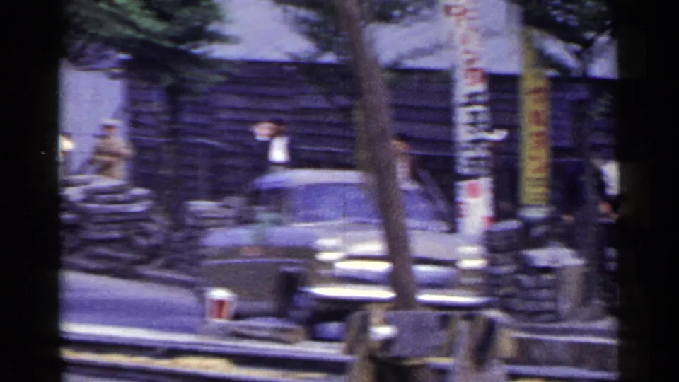 Vintage car and pedestrians on a Japanese city street beside sandbags, construction barriers, trees, and vertical signage.