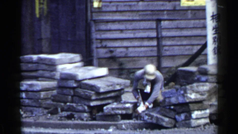In 1953 Japan, a capped worker crouches at a street construction site, hammering stone blocks beside a wooden building and signage.