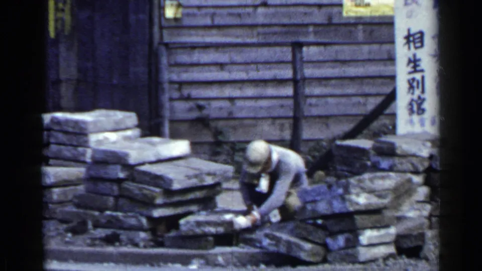 A 1953 Japan street view shows a worker using a hammer on stone slabs, with stacked blocks and a wooden facade behind him.
