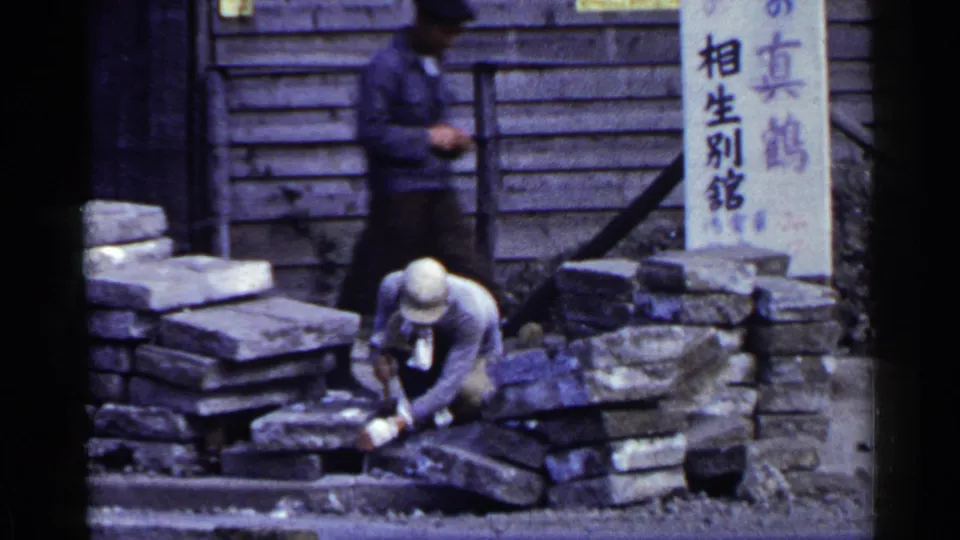 A 1953 Japan construction scene shows a crouching stone worker in the foreground and a passerby near a wooden building with Japanese signage.