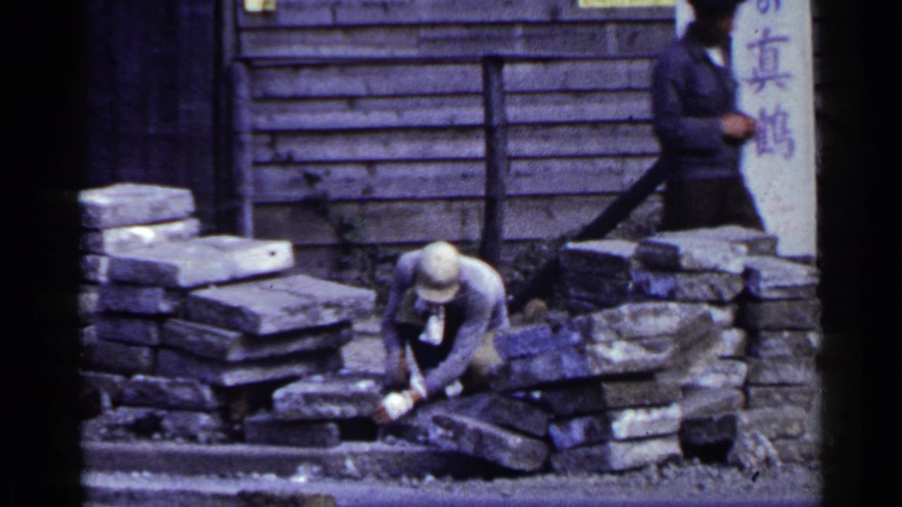 Man crouching among stone blocks outside a wooden building with Japanese signs.
