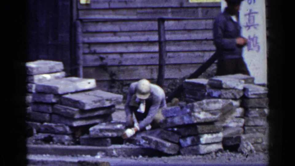 In Japan, 1953, a man in a light hat crouches to handle stone blocks near a wooden facade with vertical Japanese signage.