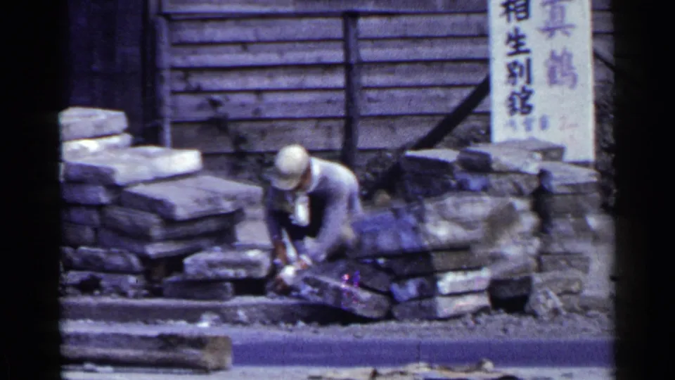 In Japan, 1953, the worker remains crouched beside uneven stone blocks, with wooden siding and Japanese signs behind him.