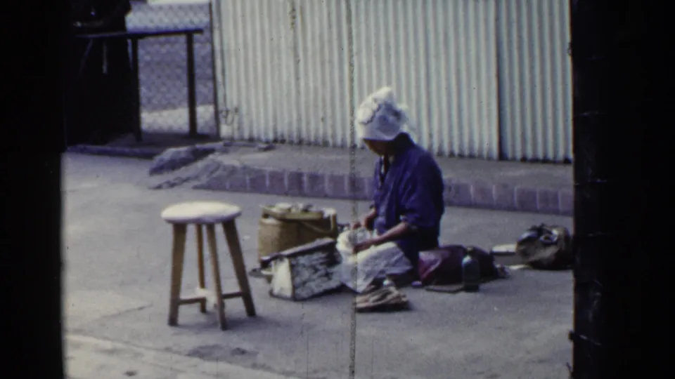 In Japan, 1953, a woman in a white head covering sits on pavement beside a stool, woven basket, and goods or tools.