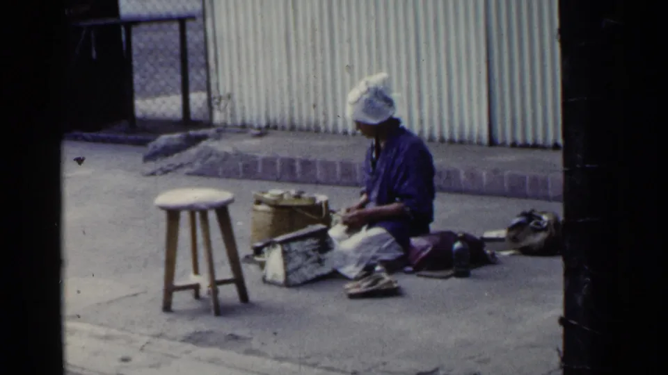 In Japan, 1953, a woman seated outdoors works with items near a small stool, basket, bag, and corrugated wall.