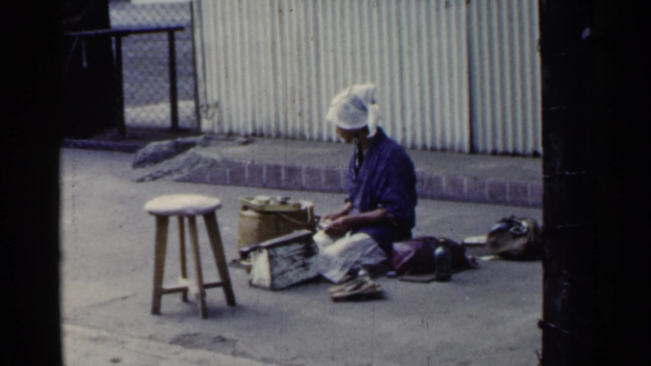 Woman seated on a street beside baskets, tools, and a wooden stool.