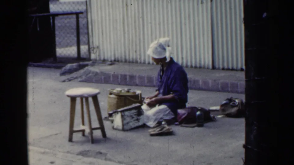 In Japan home-movie footage from 1953, a woman sits on pavement beside baskets, tools, a stool, and corrugated siding.