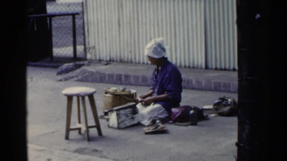 The woman remains seated in a 1953 Japan street scene, focused on hand work beside a basket, tools, bags, and a stool.