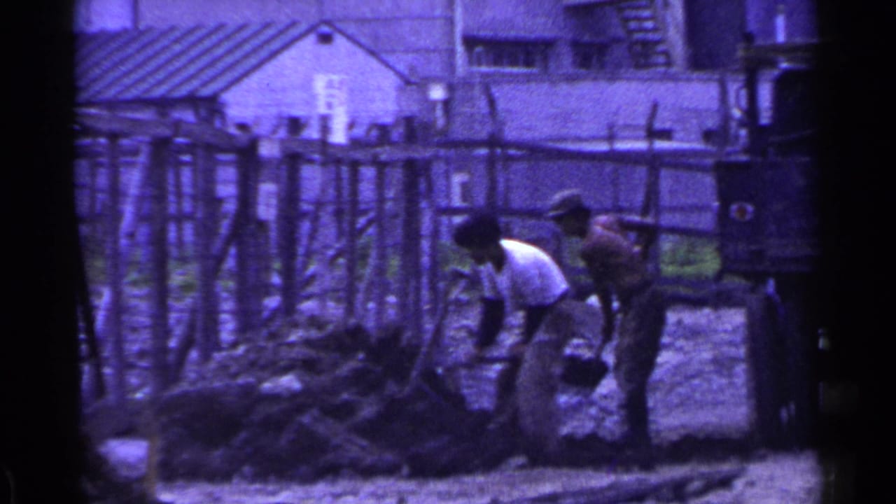 Two workers continue shoveling near a rough fence with buildings behind them.