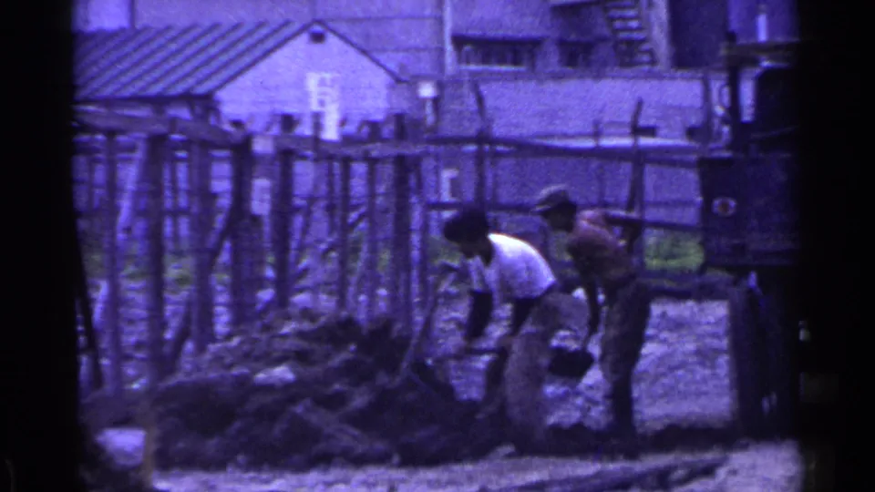 Two adults work with shovels beside a soil heap in an outdoor Japanese setting, with fencing and simple structures behind them.