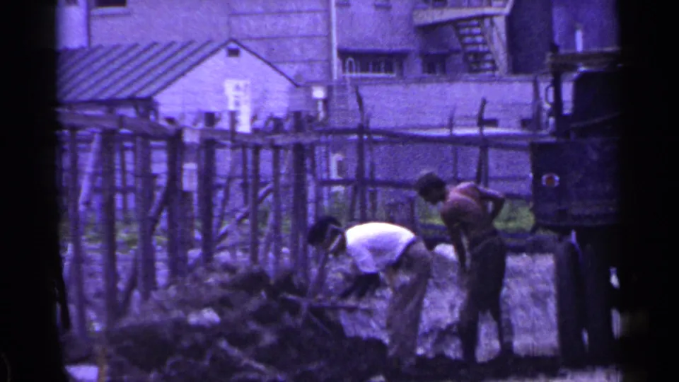 A worker bends toward the ground with a shovel while another stands near the soil pile, framed by fencing and buildings in Japan.