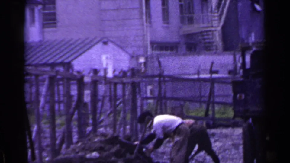 A worker bends low over a mound of earth in 1953 Japan footage, with rough fencing and modest buildings visible behind the work area.