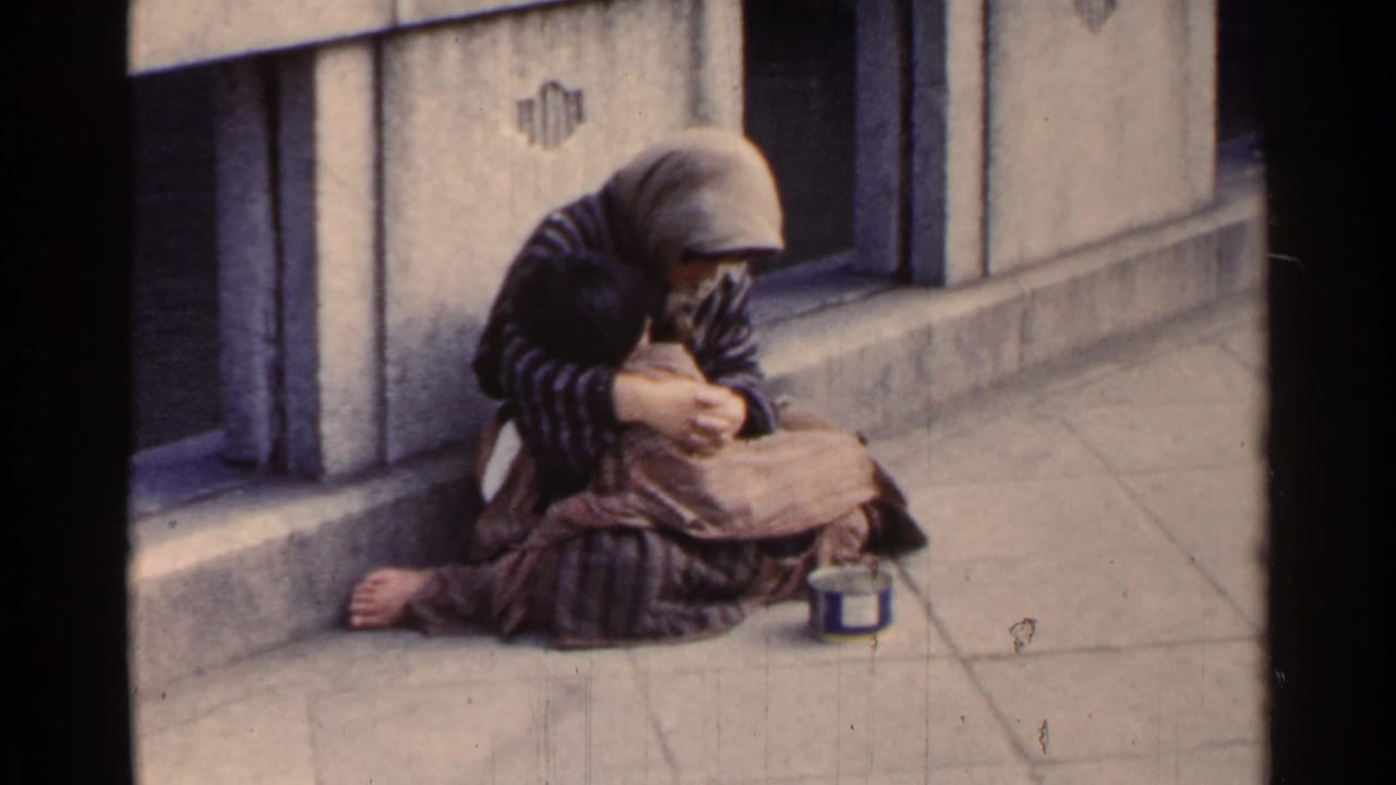Huddled person sits on a bridge sidewalk with a small tin can nearby.