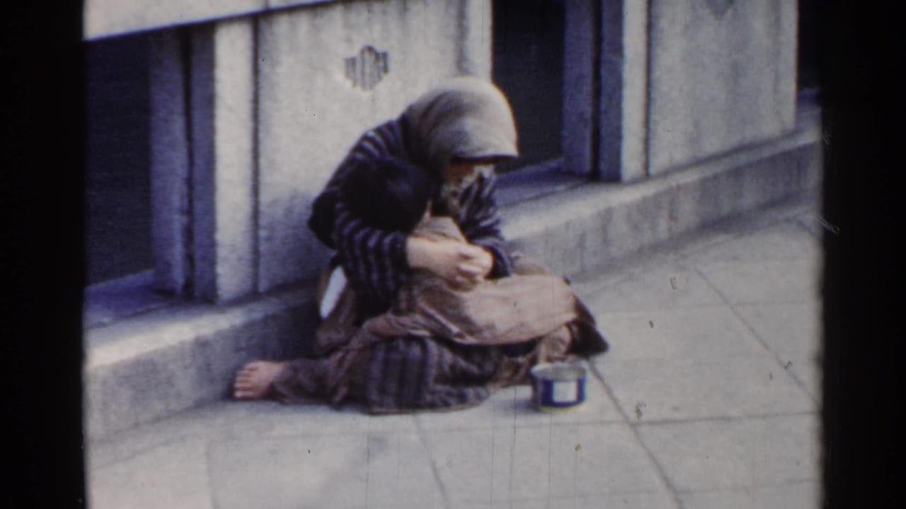 Person wrapped in clothing sits by a concrete bridge rail with a tin can nearby.
