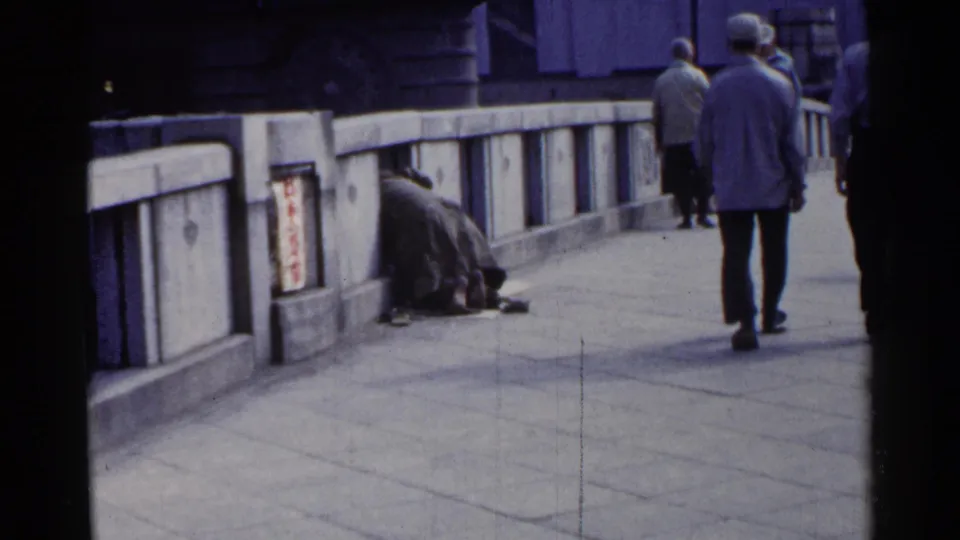 A wider 1953 Japan street view shows the huddled person beside the bridge rail as pedestrians walk past in the background.