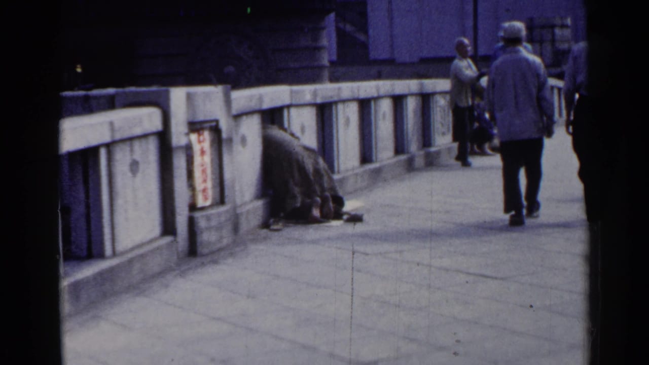 Pedestrians walk along a concrete bridge while a huddled person sits beside the railing.