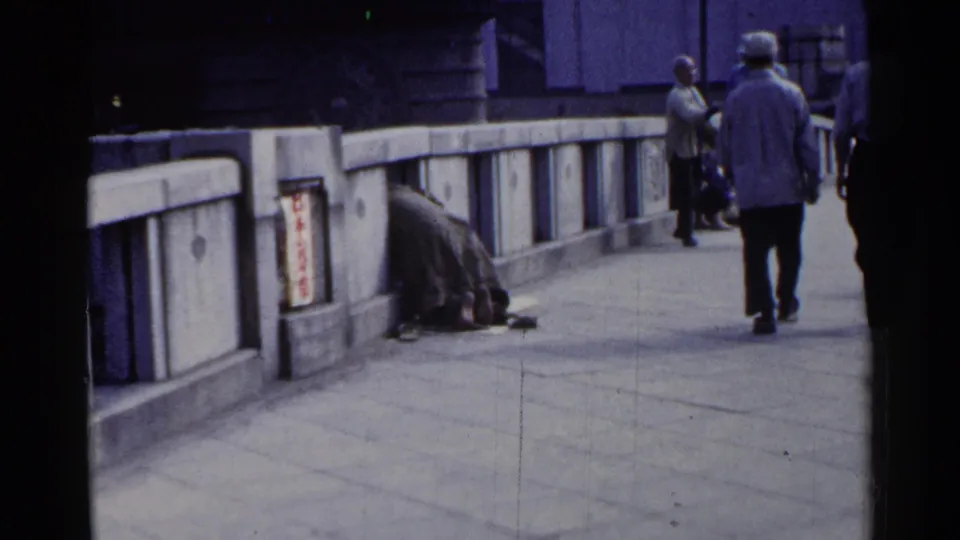 1953 Japan 8mm frame showing pedestrians on a concrete bridge walkway and a huddled person near a red-and-white Japanese sign.