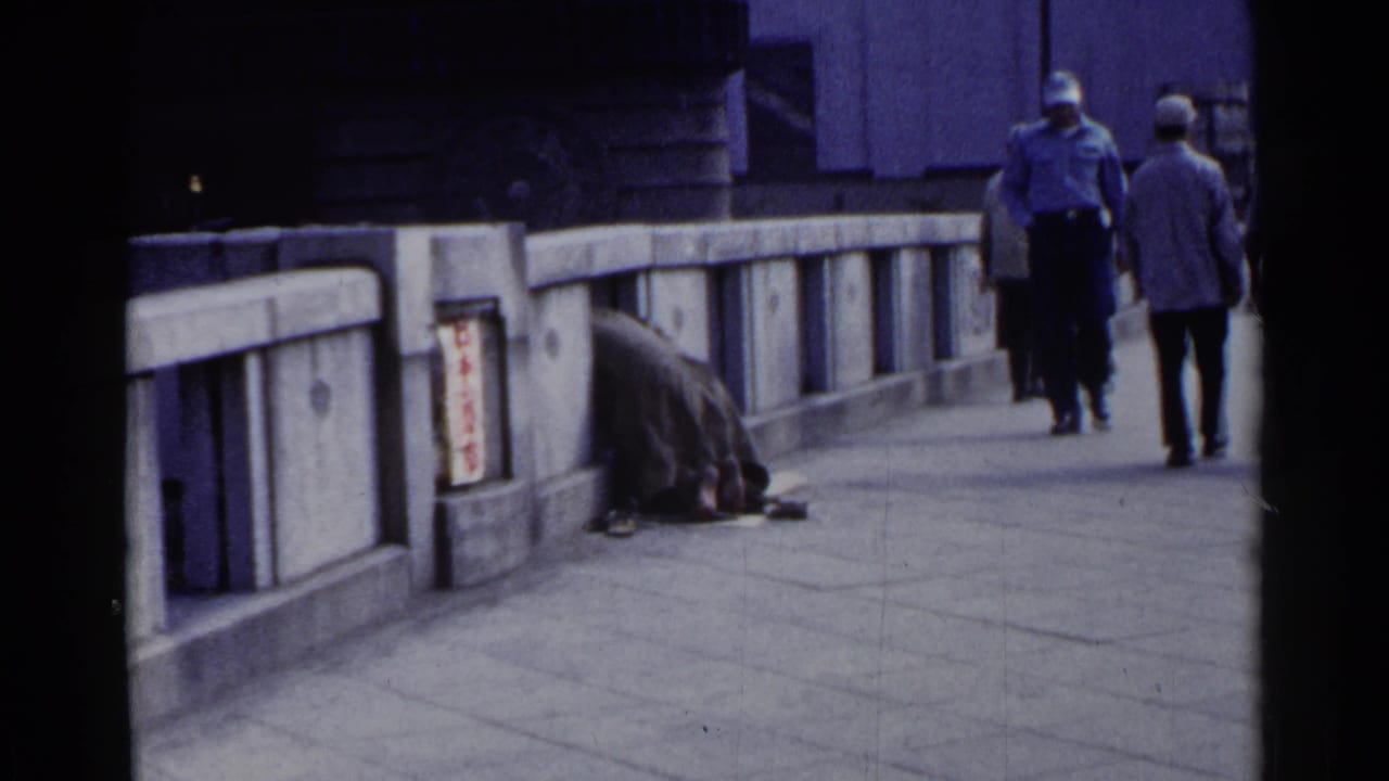 Two pedestrians walk away on a bridge as a huddled person remains beside the railing.