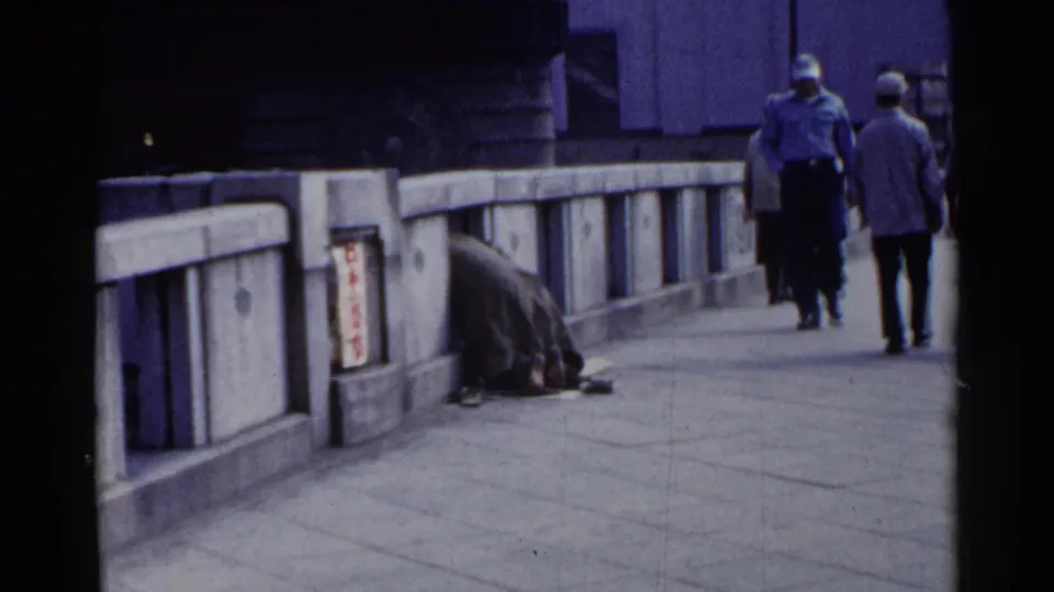 1953 Japan street footage frame with pedestrians moving along a bridge walkway past a person huddled by the concrete railing.