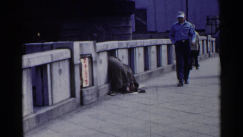 1953 Japan 8mm frame showing a man in a cap and light shirt walking toward the camera on a bridge beside a huddled person.