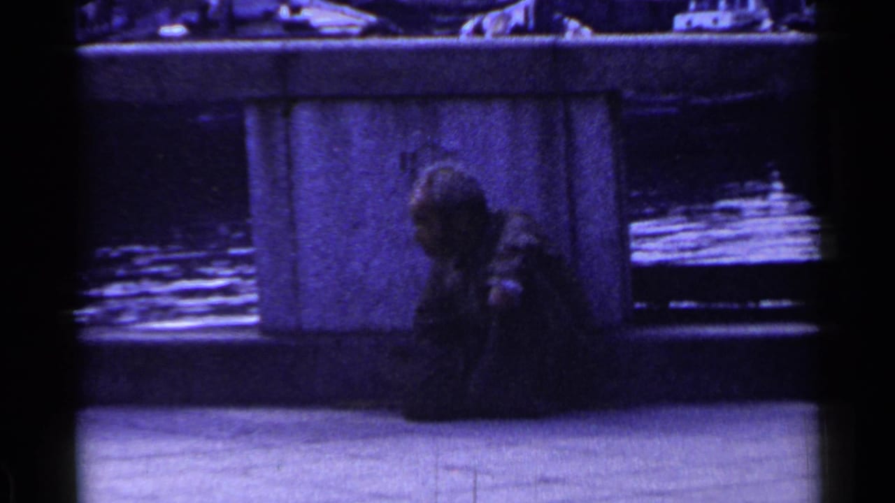 Man seated beside a concrete bridge pillar with water and blurred traffic behind him.