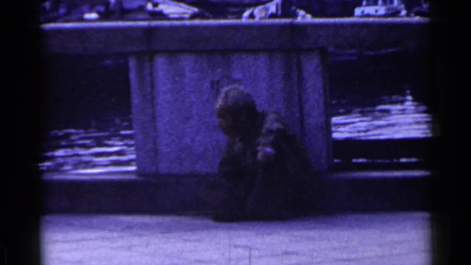1953 Japan 8mm frame showing a man sitting low beside a concrete bridge pillar above reflective water as traffic passes behind.