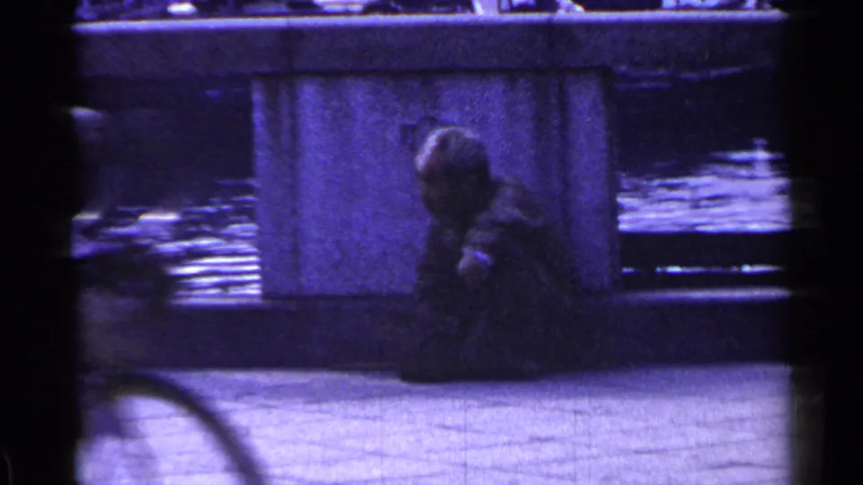 1953 Japan home-movie frame with a seated man by a bridge pillar and motion-blurred traffic crossing the scene.