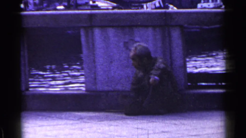 1953 Japan 8mm frame of a man seated on pavement by a concrete bridge structure, with water reflecting light behind him.