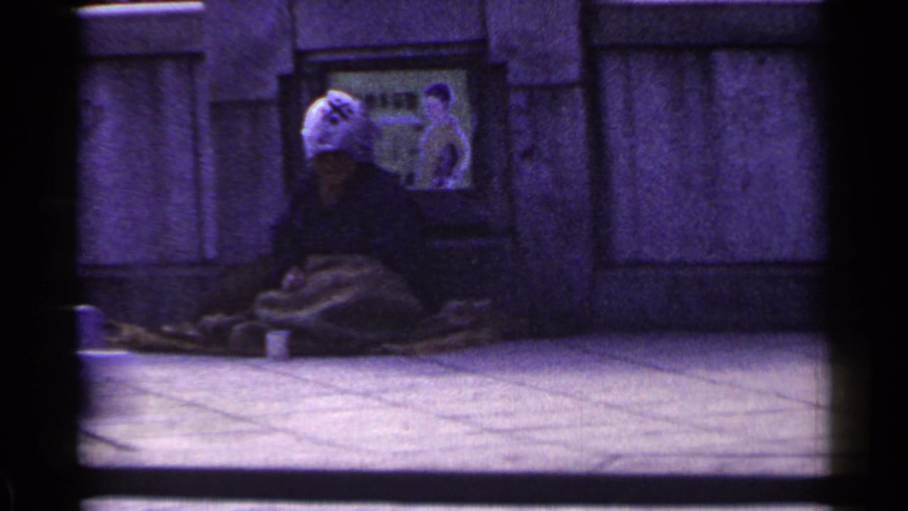 Bundled person in a light hat sits against a wall while a pedestrian passes on a city sidewalk.