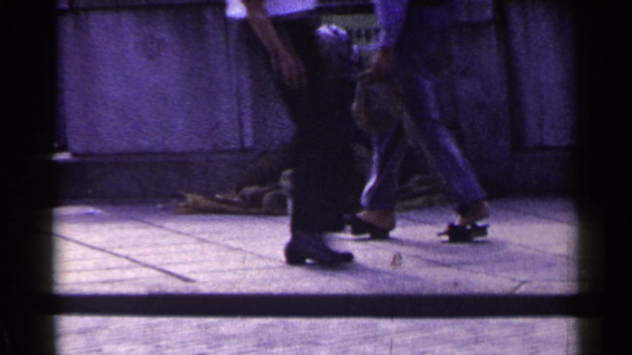 Pedestrians pass near the seated person, with legs and shoes prominent beside the wall.