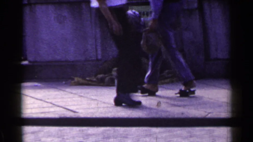 1953 Japan sidewalk frame showing passing pedestrians near a bundled person seated along a concrete wall.